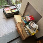 マンション外廊下に置かれた本入り段ボールとカゴ、買取前にまとめられた書籍の山。
