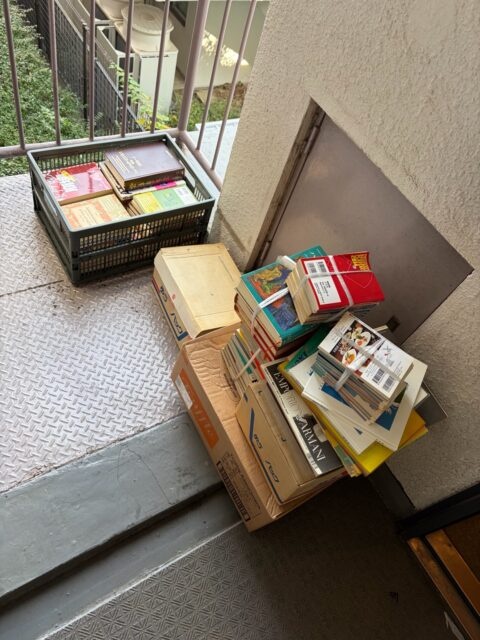 マンション外廊下に置かれた本入り段ボールとカゴ、買取前にまとめられた書籍の山。
