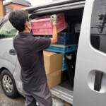 黒いポロシャツの男性が、折りたたみコンテナに入った書籍をワゴン車へ積み込んでいる様子。